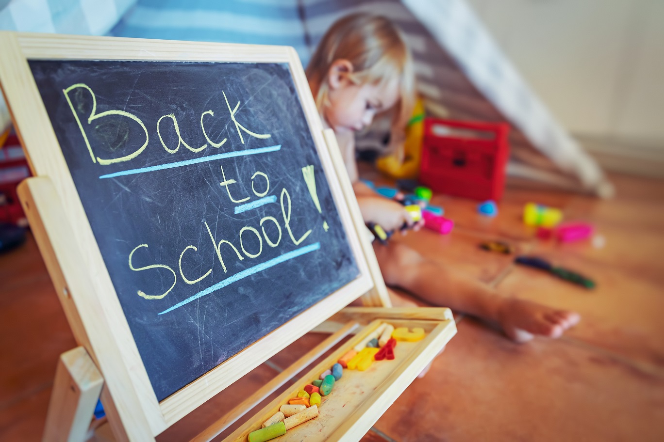 Back to school, pretty child with pleasure studying and playing at the same room at home, a little chalkboard with writing words on it standing among different toys in a baby room